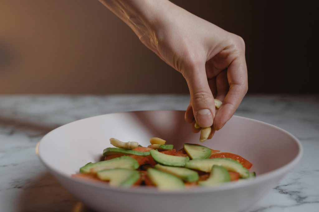 Hand adding cashews to a fresh vegan salad with avocado slices in a ceramic bowl.